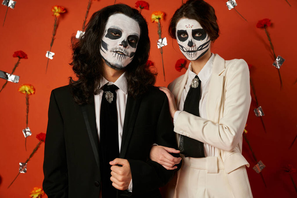 couple in eerie sugar skull makeup and festive attire looking at camera on red backdrop with flowers - Photo, Image