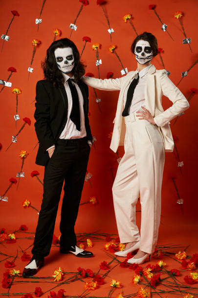 longitud completa de la pareja con estilo en el maquillaje del cráneo y trajes de pie sobre fondo rojo con claveles - Foto, imagen