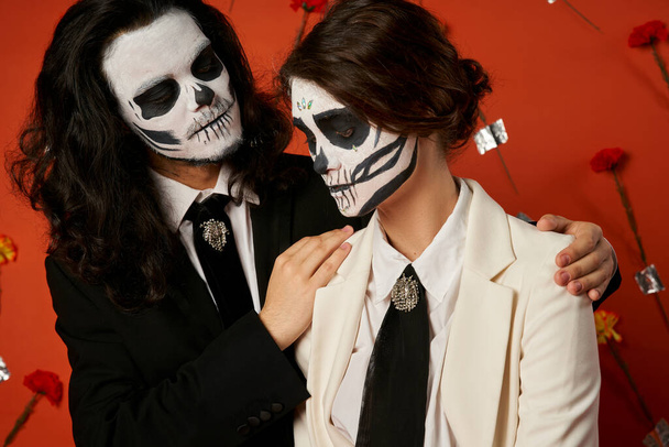 spooky man embracing shoulders of woman in suit, dia de los muertos couple on red floral backdrop - Photo, Image