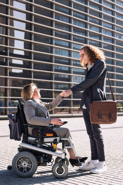 business man and woman using wheelchair smiling while shaking hands in front of office building in financial district, concept of diversity and urban lifestyle, copy space for text - Фото, изображение