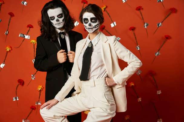 couple in catrina makeup and elegant attire posing on red background with flowers, Day of Dead - Photo, Image