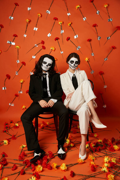 Stilvolles Paar in Totenkopf-Make-up und Anzügen posiert auf Stühlen vor rotem Hintergrund mit Blumen, Day of Dead - Foto, Bild