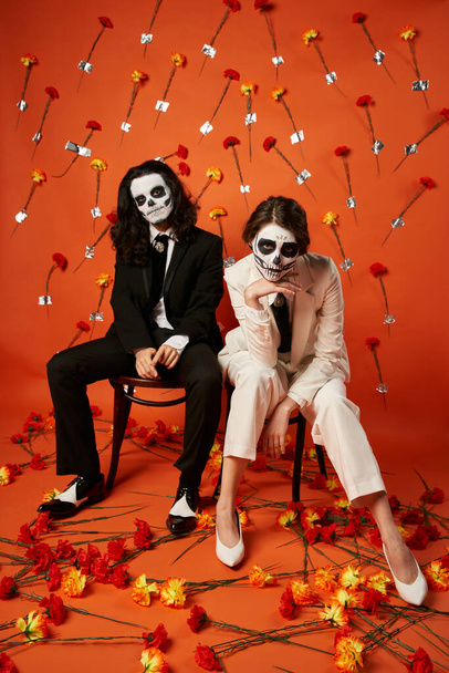 trendy couple in skull makeup and suits looking at camera on chairs on red backdrop with carnations - Photo, Image