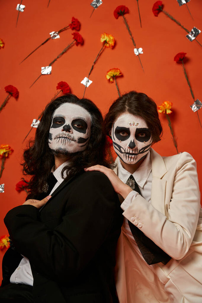 Gruseliges Paar in dia de los muertos Make-up und festlicher Kleidung auf rotem Hintergrund mit Blumen - Foto, Bild