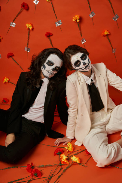 couple in scary catrina makeup and suits sitting on floor in red studio with carnation flowers - Foto, Bild