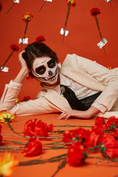 Elegante Frau in Zuckerschädel-Make-up und weißem Anzug im roten Studio neben Nelken liegend - Foto, Bild