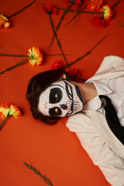 γυναίκα σε dia de los muertos μακιγιάζ και κοστούμι ξαπλωμένη σε κόκκινο φόντο με λουλούδια, top view - Φωτογραφία, εικόνα