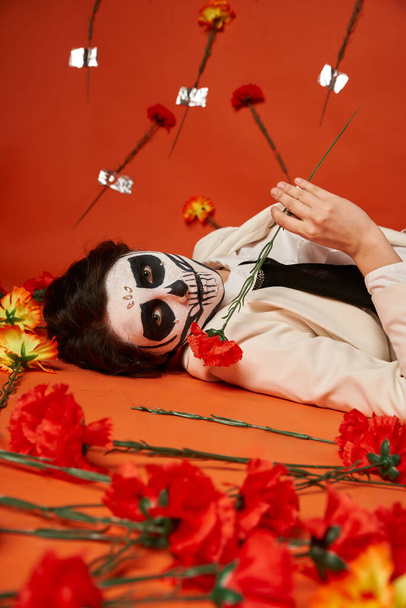 woman in sugar skull makeup and suit lying down and looking at camera near carnations in red studio - Photo, Image