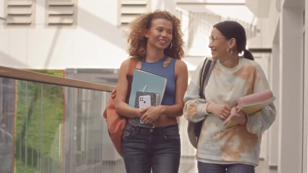 Střední zpomalení dvou krásných asijských a kavkazských přítelkyň kráčejících po chodbě vysoké školy s konverzací - Záběry, video
