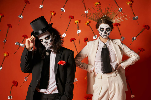 fiesta día de los muertos, pareja en maquillaje catrina y atuendo festivo en estudio rojo con flores - Foto, imagen