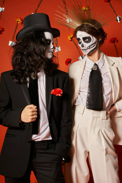fiesta de día de los muertos, pareja en maquillaje aterrador mirándose en estudio rojo con flores - Foto, Imagen