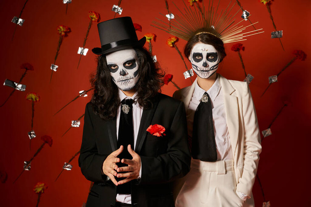 día de los muertos fiesta, pareja en maquillaje de miedo mirando a la cámara en estudio rojo con claveles - Foto, imagen