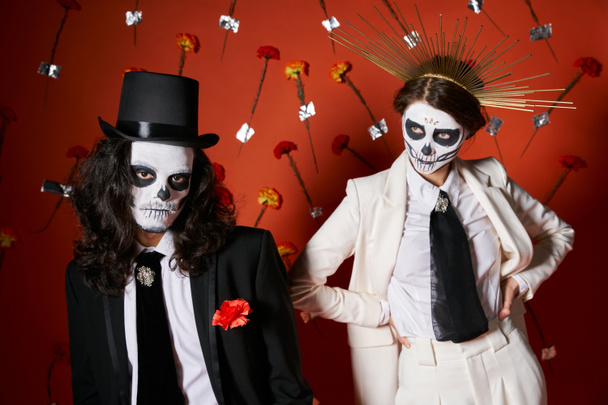 pareja en día de los muertos cráneo maquillaje y elegante atuendo posando sobre fondo rojo con claveles - Foto, imagen