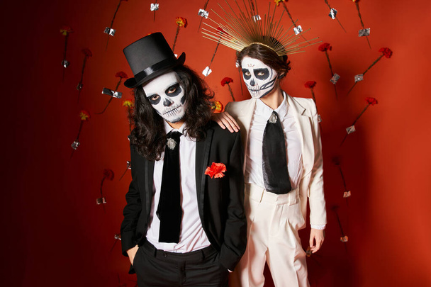 Elegantes Paar in dia de los muertos Totenkopf-Make-up blickt in die Kamera auf rotem Hintergrund mit Blumen - Foto, Bild