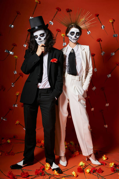 full length of festive elegant couple in dia de los muertos makeup on red backdrop with floral decor - Photo, Image