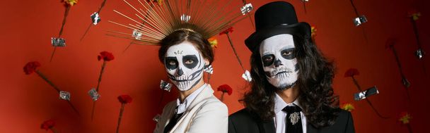 pareja en día de los muertos maquillaje y atuendo festivo posando sobre fondo rojo con flores, pancarta - Foto, imagen