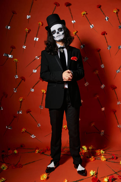 Mann in Totenkopf-Make-up und festlicher Kleidung auf rotem Blumenhintergrund, dia de los muertos - Foto, Bild
