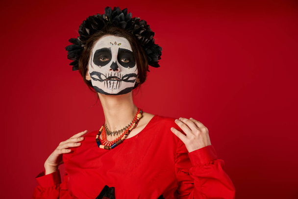 woman in traditional dia de los muertos skull makeup and colorful beads looking at camera on red - Photo, Image