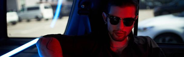 fiatal jóképű férfi napszemüvegben tetovált karon pózol az autójában, divat koncepció, banner - Fotó, kép