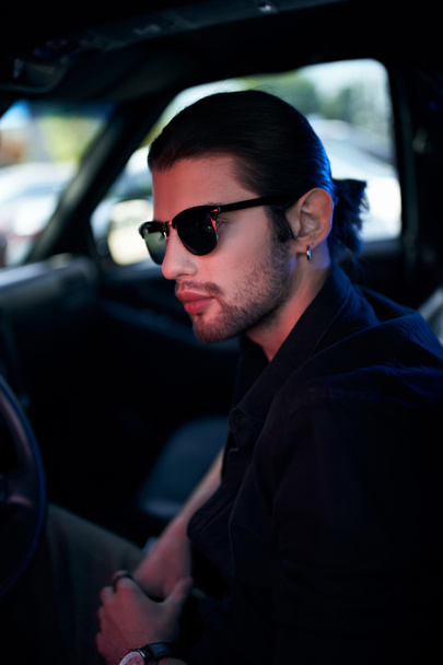 colpo verticale di bel giovane uomo con orecchino e occhiali da sole in posa nella sua auto, concetto di moda - Foto, immagini