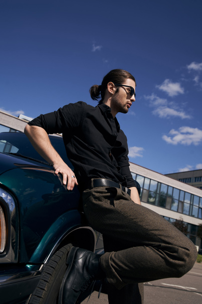 сексуальний стильний чоловік з бородою і хвостом позує біля своєї зеленої машини і дивиться в сторону, концепція моди - Фото, зображення
