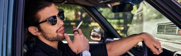 młody brodaty mężczyzna z zegarkiem na rękę i okularami przeciwsłonecznymi palący papierosa podczas pozowania w samochodzie, baner - Zdjęcie, obraz