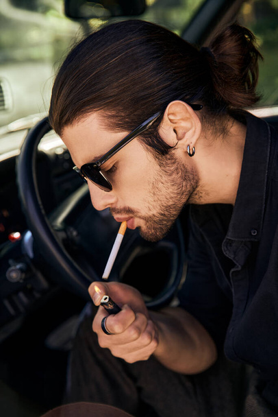 Arabasında sigara yakan küpe ve güneş gözlüklü yakışıklı seksi erkek manken, seksi şoför. - Fotoğraf, Görsel