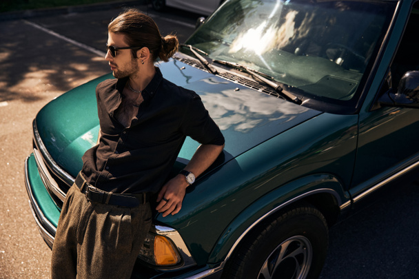 affascinante giovane uomo con accessori alla moda appoggiato leggermente sulla sua auto e guardando altrove, moda - Foto, immagini