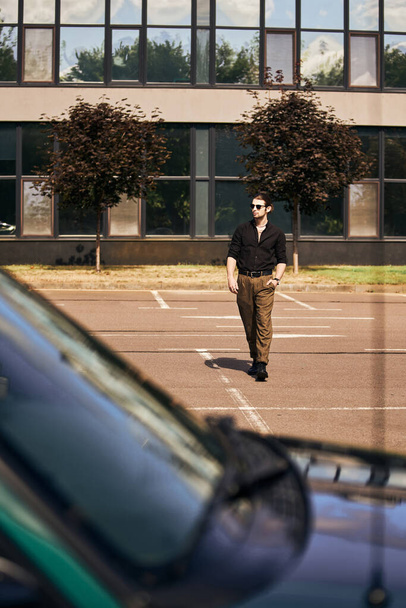όμορφος νεαρός άνδρας σε μαύρο μοντέρνα ενδυμασία με τα πόδια στο αυτοκίνητό του με το χέρι στην τσέπη, έννοια της μόδας - Φωτογραφία, εικόνα