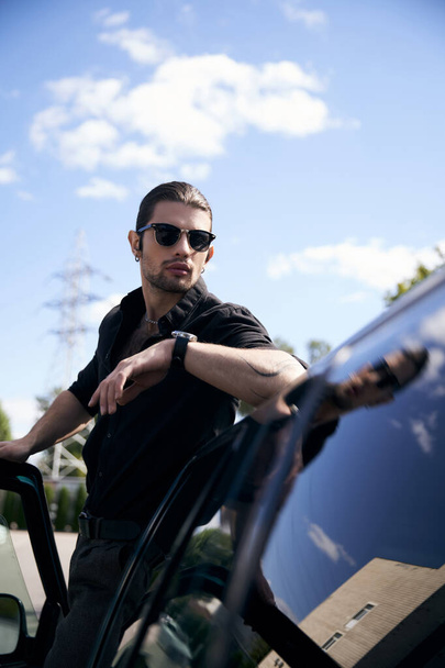 Güneş gözlüklü ve dövmeli yakışıklı bir erkek model. Arabasında koluyla dışarıda poz veriyor. Seksi şoför. - Fotoğraf, Görsel