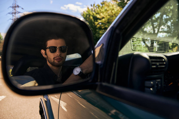 verleidelijke elegante man met zonnebril in zwarte stijlvolle outfit op zoek in zijspiegel, sexy chauffeur - Foto, afbeelding