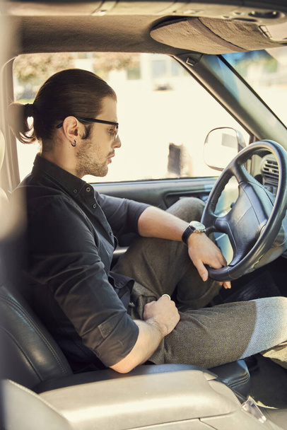 pensive stijlvol mannelijk model met accessoires in zwarte outfit achter stuurwiel, sexy driver - Foto, afbeelding
