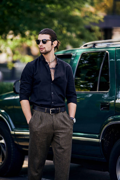 сексуальна людина з аксесуарами в стильному вбранні, що стоїть поруч зі своєю машиною з руками в кишенях - Фото, зображення