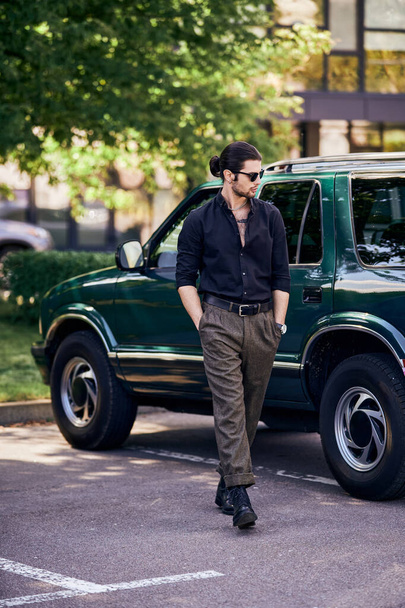 przystojny sexy mężczyzna w okulary przeciwsłoneczne w czarny koszula chodzenie w pobliżu jego samochód i patrząc daleko, moda - Zdjęcie, obraz