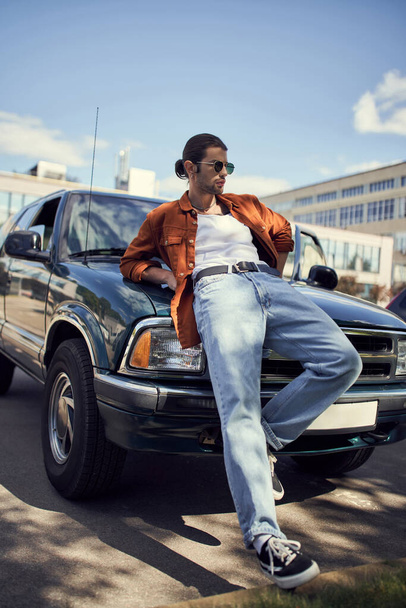 goed uitziende sexy man in levendige stijlvolle kleding leunend op zijn auto weg te kijken, mode en stijl - Foto, afbeelding