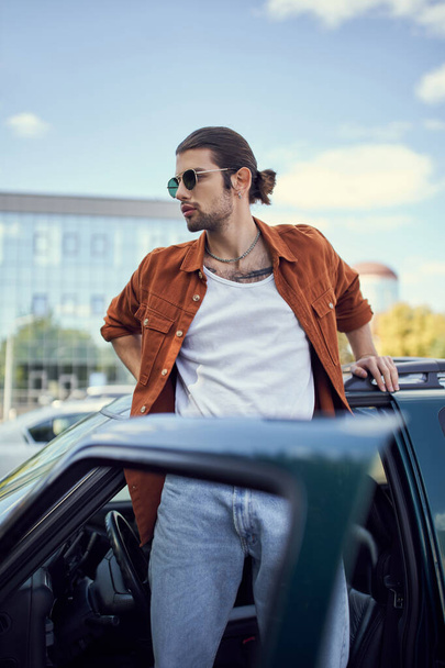 jovem modelo masculino encantador posando em traje vibrante ao lado de seu carro com porta aberta, conceito de estilo - Foto, Imagem