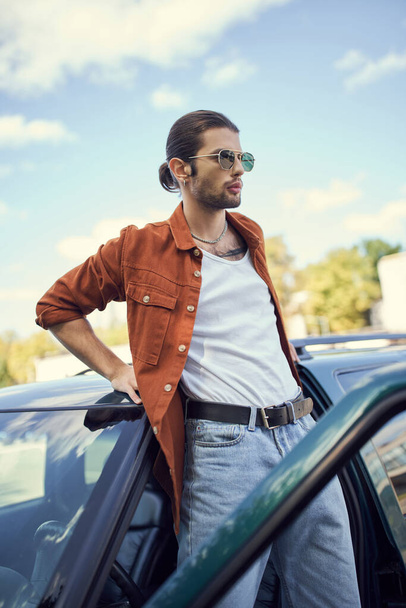 encantador modelo masculino joven en camisa marrón y vaqueros azules de pie junto a su coche y mirando hacia otro lado - Foto, imagen