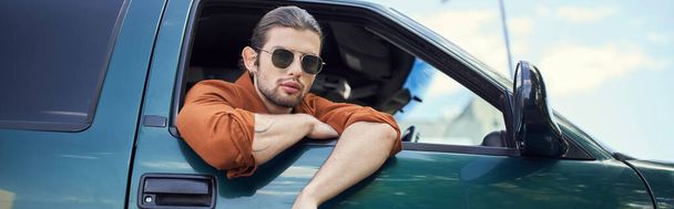 Güneş gözlüklü çekici bir adam arabanın camından kameraya, moda konseptine, afişe bakıyor. - Fotoğraf, Görsel