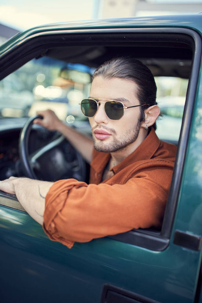 appealing young man with earring in trendy brown shirt with sunglasses looking out of car window - Photo, Image