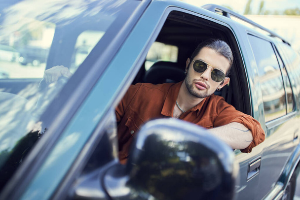 handsome young man in trendy outfit with accessories looking out of car window, sexy driver - Фото, изображение