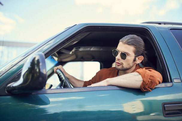 appealing young man with ponytail and earring in brown shirt looking out of car window, sexy driver - Photo, Image