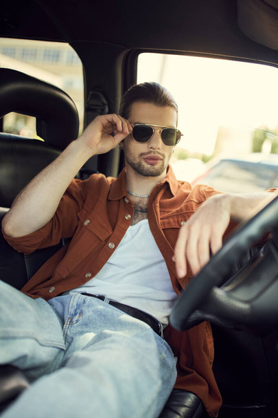 Şık giyinmiş yakışıklı genç adam arabada dinlenirken güneş gözlüğüne dokunuyor, seksi şoför. - Fotoğraf, Görsel