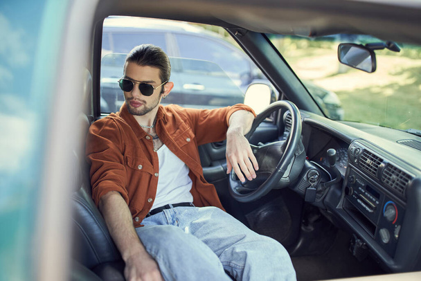 красивый мужчина в модной коричневой рубашке и джинсах расслабляется в машине за рулем, мода - Фото, изображение