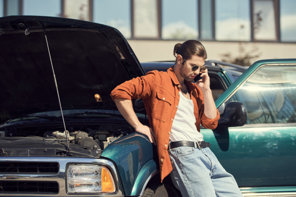 przystojny kierowca z okularami przeciwsłonecznymi w brązowej koszuli stojący obok samochodu dzwoniący do ubezpieczyciela - Zdjęcie, obraz
