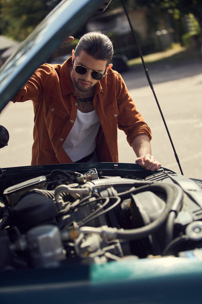 ελκυστικός νεαρός άνδρας σε καφέ πουκάμισο με αξεσουάρ κοιτάζοντας προσεκτικά τον κινητήρα του αυτοκινήτου του - Φωτογραφία, εικόνα