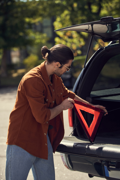 vertical shot of stylish man with sunglasses putting warning triangle in his car trunk, sexy driver - Фото, изображение