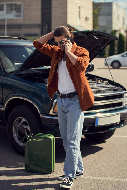 knappe man met zonnebril en paardenstaart aan de telefoon met zijn verzekeraar, benzinetank - Foto, afbeelding