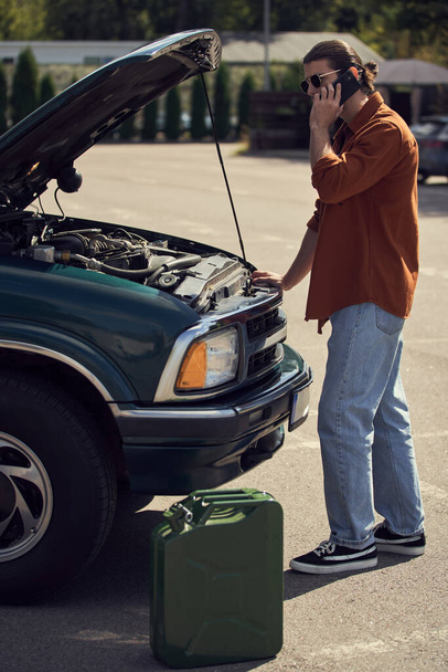 вертикальный снимок стильного мужчины, стоящего рядом с автомобилем и канистрой с бензином, разговаривающего по телефону со страховщиком - Фото, изображение