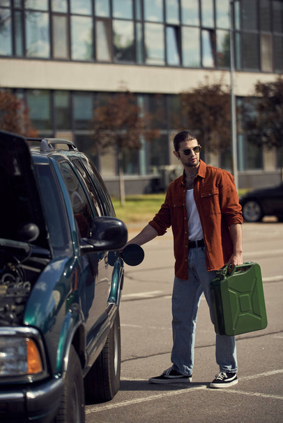 красивая сексуальная мужская модель в коричневой рубашке и джинсах с элегантными солнцезащитными очками для заправки своего автомобиля - Фото, изображение