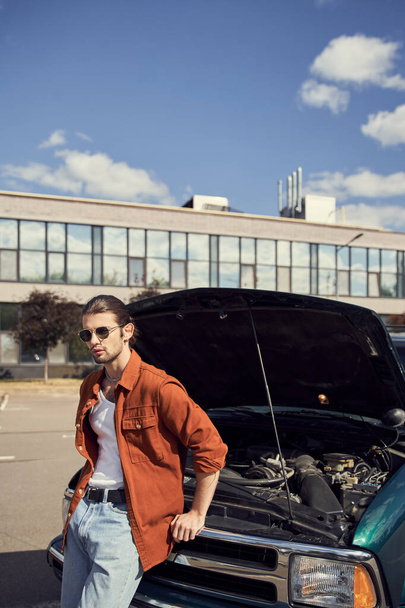 pionowe ujęcie atrakcyjnego eleganckiego mężczyzny z eleganckim wyglądem stojącego w pobliżu samochodu z otwartą maską silnika - Zdjęcie, obraz
